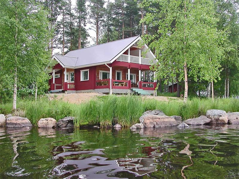 Punainen hirsitalo katettuine terasseineen järven rannalla koivujen ja mäntyjen keskellä