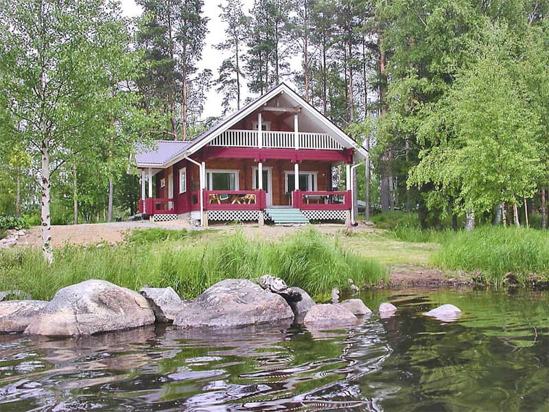 Punainen hirsitalo parvekkeineen Käpyniemen rannalla, edessä kiviä ja järven vesi